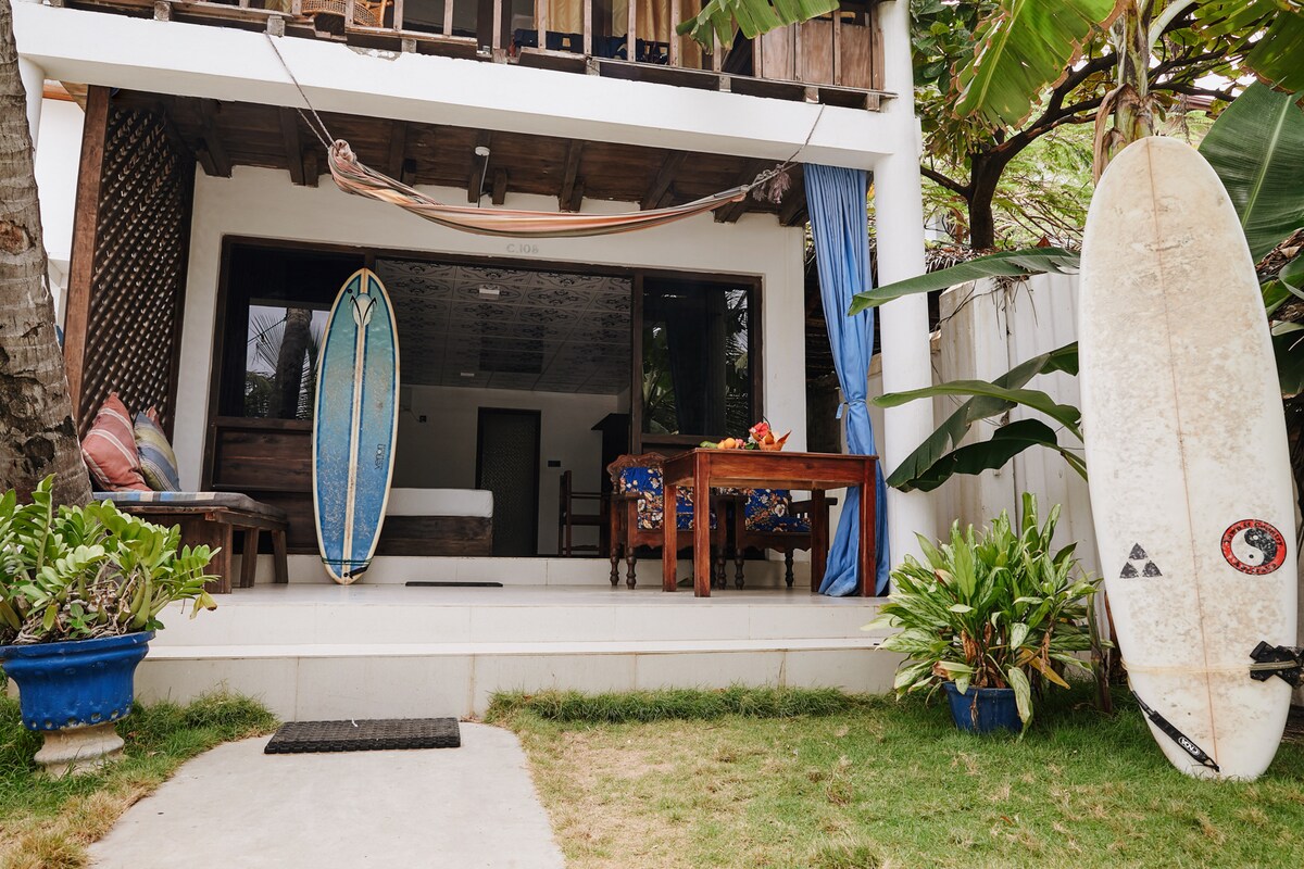 A welcoming outdoor area features a well-maintained lawn and potted plants. Two surfboards are placed near a seating area with a wooden table and colorful chairs. The entrance to the room is framed by a spacious window showcasing views of the interior.