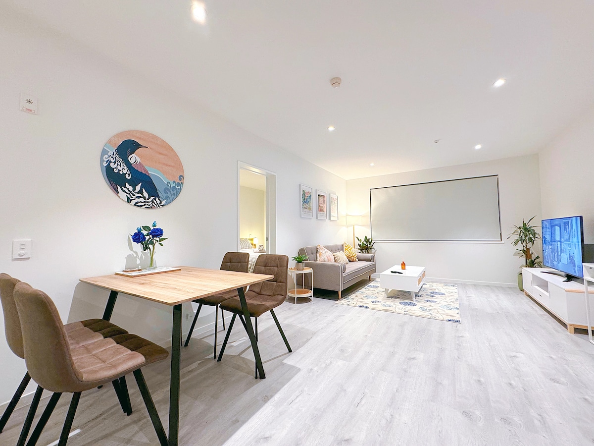An open living area is highlighted by a wooden dining table set for four, accompanied by plush brown chairs. A stylish grey sofa is positioned against the wall, complemented by a television on a low white unit. Decorative plants and wall art add personality to the space.