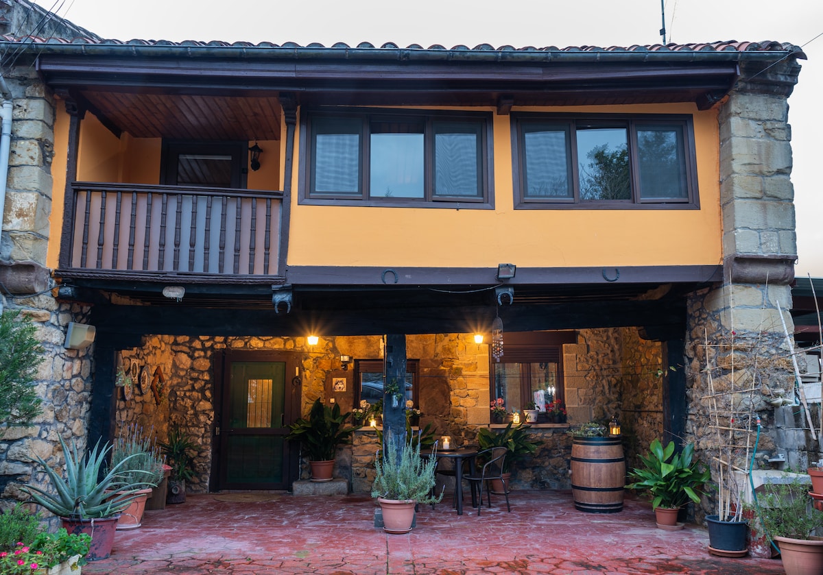 The exterior of the two-story house showcases a rustic design with stone walls and wooden beams. The entrance is framed by potted plants and features an inviting porch area with seating. Large windows provide a view of the cozy interior, illuminated by warm lighting.