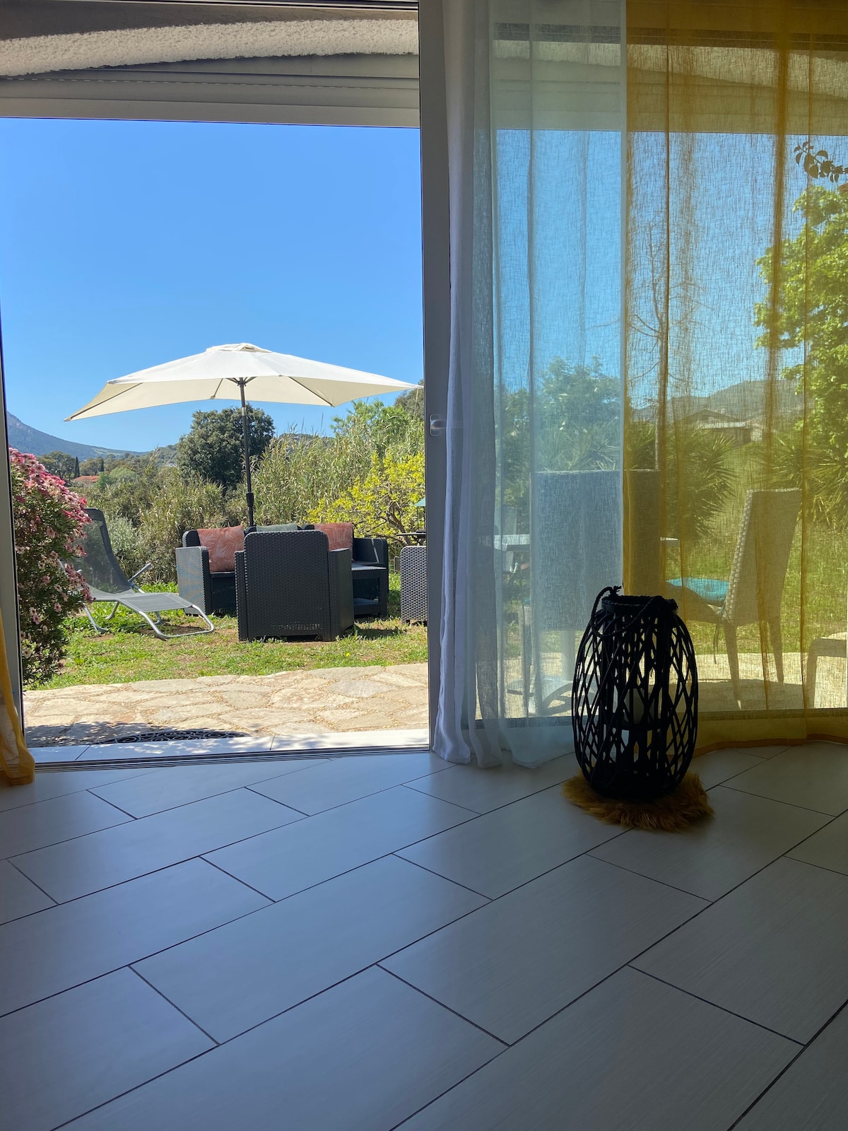 A view from inside the home opens to a garden area featuring an umbrella and seating arrangement. Sunlight illuminates the space, highlighting the outdoor greenery and surrounding mountains. A decorative lantern adds a touch of style near the open door.