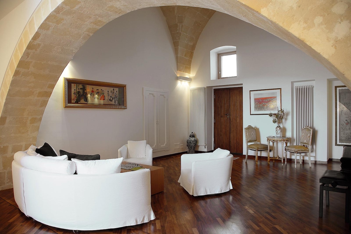 A spacious living area is highlighted by a curved stone ceiling and warm wooden flooring. Two white, modern sofas face each other, complementing a low wooden coffee table. Decorative art is displayed on the wall, while elegant chairs and a door add character to the room.