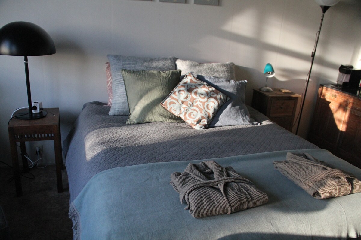 A well-appointed sleeping area features a spacious bed with a textured blanket and a variety of decorative pillows. Soft natural light streams in, highlighting the bedside lamp and wooden nightstand. Towels are neatly arranged on the bed, adding a touch of accommodation for guests.