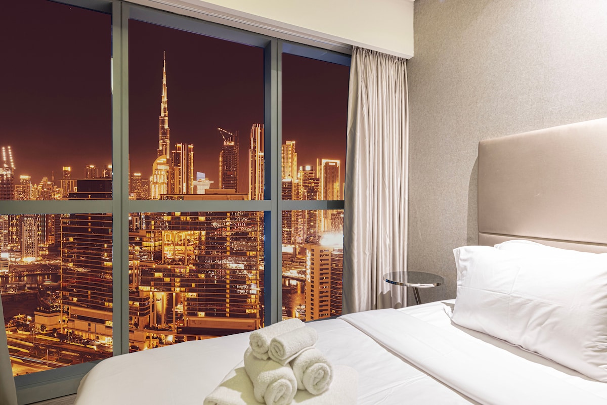 A stylish bedroom with a spacious bed, accented by neatly arranged towels. Floor-to-ceiling windows provide a striking view of the illuminated city skyline, including the Burj Khalifa, enhancing the room's ambiance.
