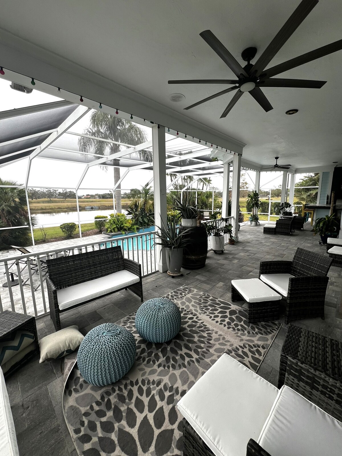 The covered outdoor patio features a spacious seating area with dark wicker chairs and light cushions. Two textured blue poufs are placed on a patterned area rug, while ceiling fans provide comfort. Through the mesh screens, a private pool and lush landscape are visible.