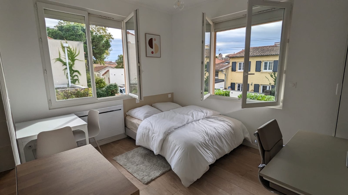 A well-lit studio space features a spacious bed dressed in crisp white linens, positioned near large windows offering views of greenery and neighborhood architecture. A simple desk and chair setup is visible, along with a cozy rug underfoot, contributing to a serene atmosphere.