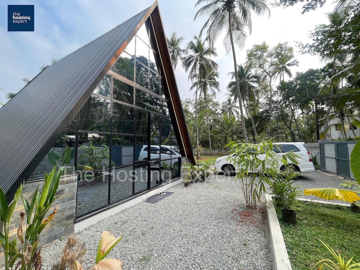 A modern A-frame structure is highlighted by large glass panels, creating a seamless connection between the indoors and the lush outdoor surroundings. The driveway is bordered by decorative gravel and tropical plants, providing a serene entrance. Palm trees sway gently in the background.