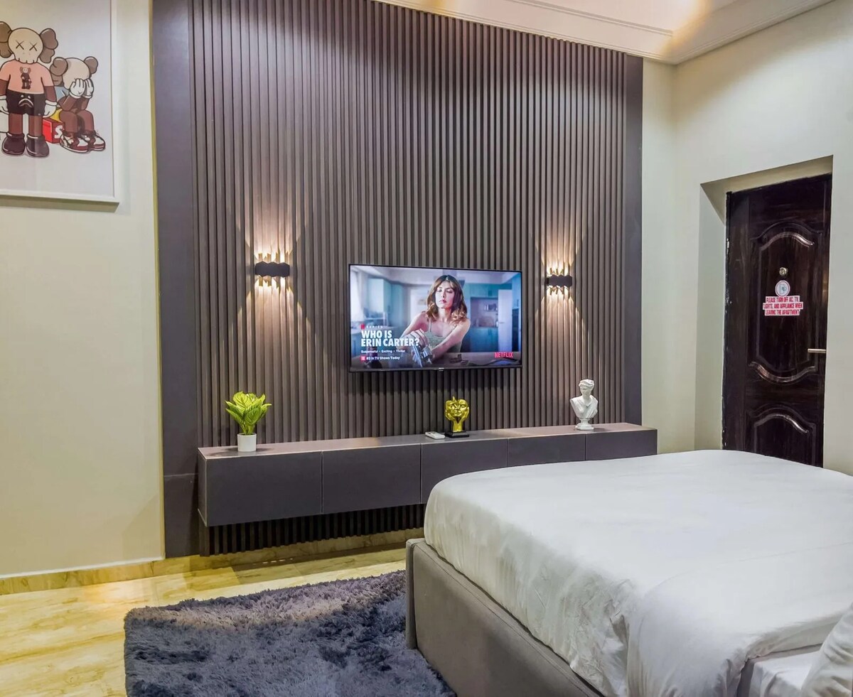 A modern bedroom is featured with a spacious bed dressed in white linens. A flat-screen TV is mounted on the wall, and a low cabinet provides storage. Two wall sconces offer soft lighting, while decorative plants add a touch of greenery.