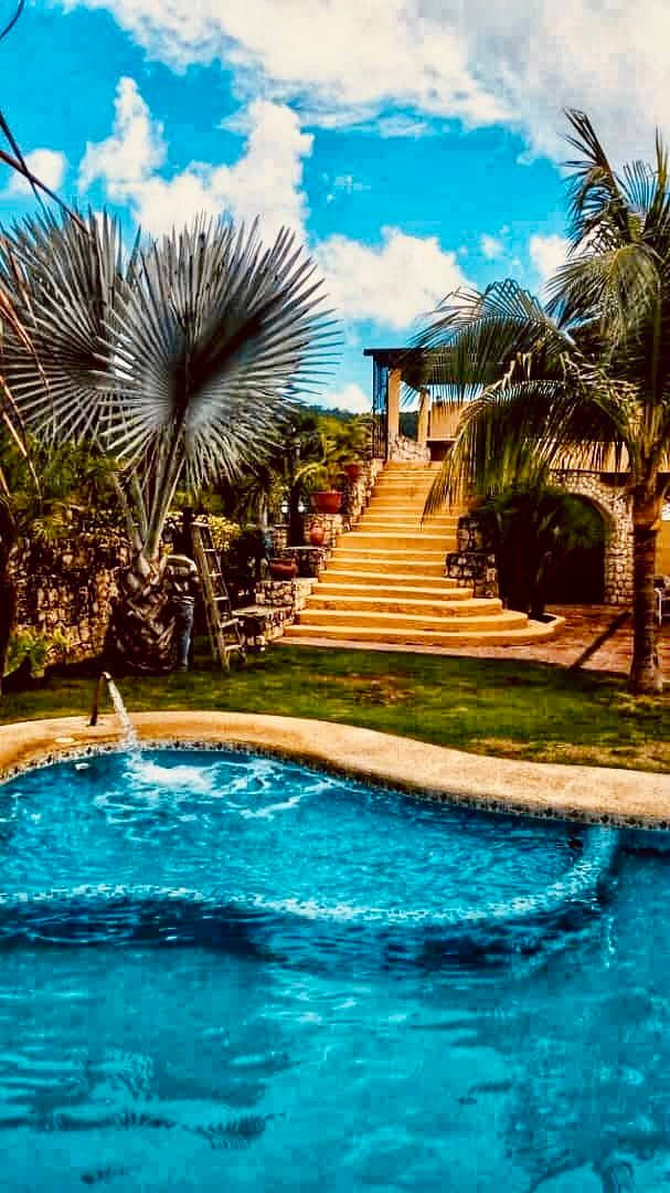A serene swimming pool is featured in the foreground, with bubbling water and vibrant blue tones. In the background, a staircase leads to a multi-level villa surrounded by lush greenery and palm trees, under a bright sky dotted with clouds.