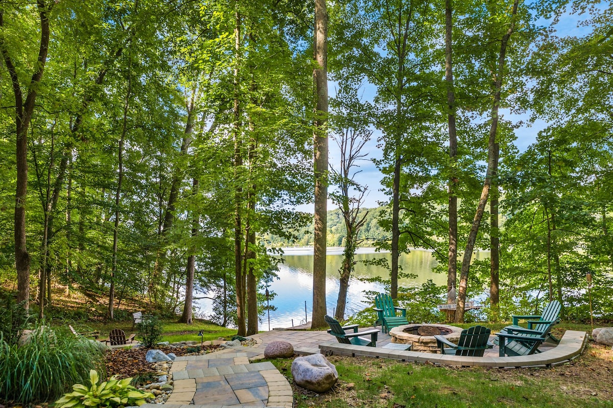 A serene outdoor space is framed by lush trees, featuring a circular stone patio with six Adirondack chairs surrounding a fire pit. The tranquil lake reflects the greenery, creating a peaceful backdrop for relaxation. The gentle slope leads to the water's edge.