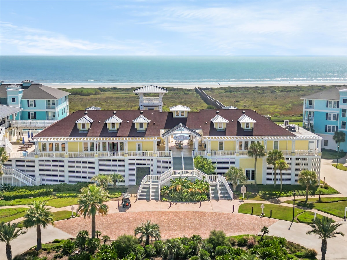 The beachfront property is presented with a bright yellow exterior and multiple gabled roofs. Palm trees are planted around the entrance pathway, leading to an inviting porch area. Beyond, the Gulf of Mexico sparkles under a clear blue sky, creating a serene coastal backdrop.