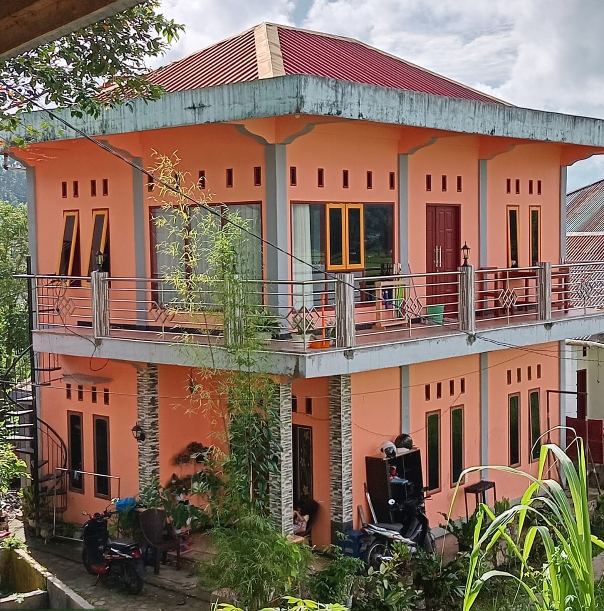 A two-story building is showcased with a vibrant orange exterior, accented by multiple windows. A balcony extends across the front, offering a view of the surrounding greenery. The entrance features double doors, and a narrow staircase is visible along the side of the structure.