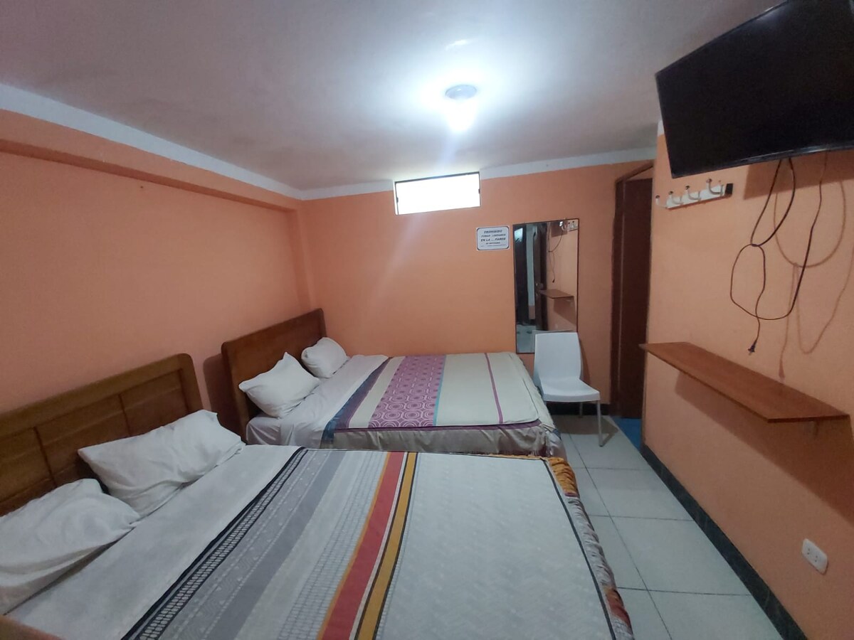 The room is arranged with two beds, each featuring neatly arranged bedding in neutral tones. A small wall-mounted television is positioned in the corner, and a wooden shelf extends along one wall. An illuminated ceiling provides soft lighting, enhancing the simple layout.