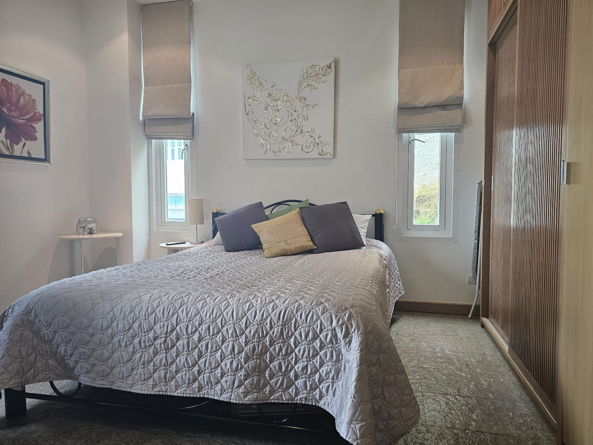 A cozy bedroom features a spacious bed adorned with a textured quilt. Two accent pillows in shades of purple add subtle color. Natural light enters through two windows, complemented by elegant window treatments. A framed floral artwork and a bedside table enhance the simple decor.