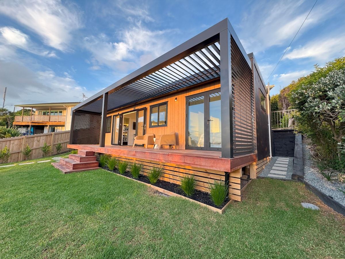 The modern beach bach is showcased with a spacious deck bordered by lush green grass. A shaded overhang provides a cozy outdoor seating area, while large windows reflect the surrounding landscape. The structure features warm wood tones and a sleek black exterior, blending with the coastal environment.