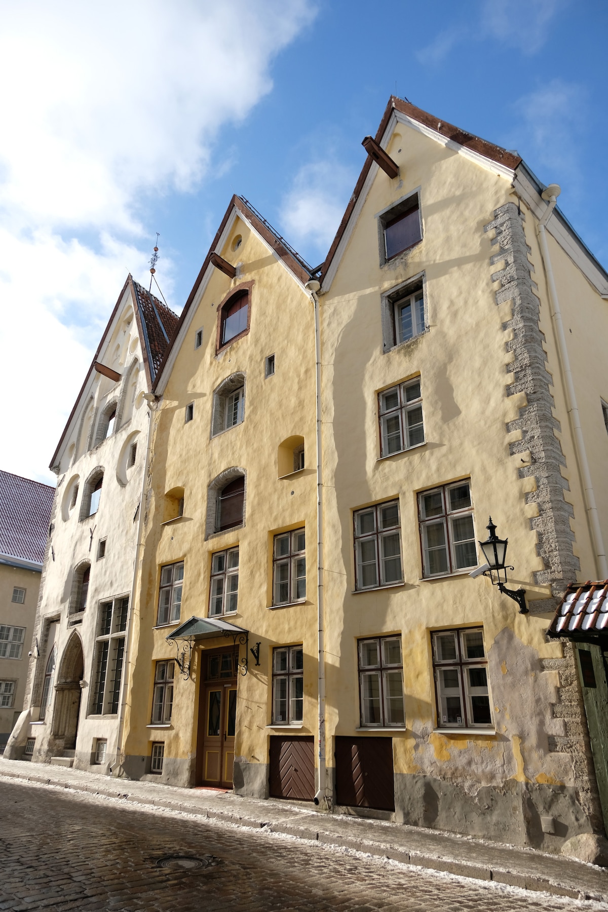 A charming historic building is displayed with a combination of yellow and gray façades. The structure features multiple windows of varying sizes, accentuated by a slate roof. The inviting entrance is framed by a small overhang and lantern, showcasing the architectural details of the landmark.