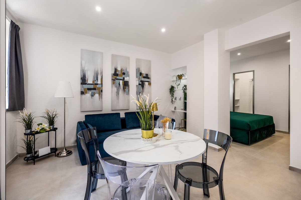 A bright living area features a white marble dining table surrounded by four transparent chairs. A dark blue sofa is positioned against the wall alongside decorative plants. Two large artworks hang above the sofa, and a doorway leads to a bedroom with a green bedspread.