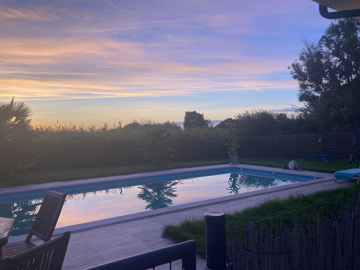 A tranquil scene is captured at dusk, featuring a clear swimming pool that reflects the colorful sky. Lush greenery surrounds the area, creating a serene atmosphere. A wooden table with chairs is positioned nearby, inviting relaxation and enjoyment of the outdoor space.