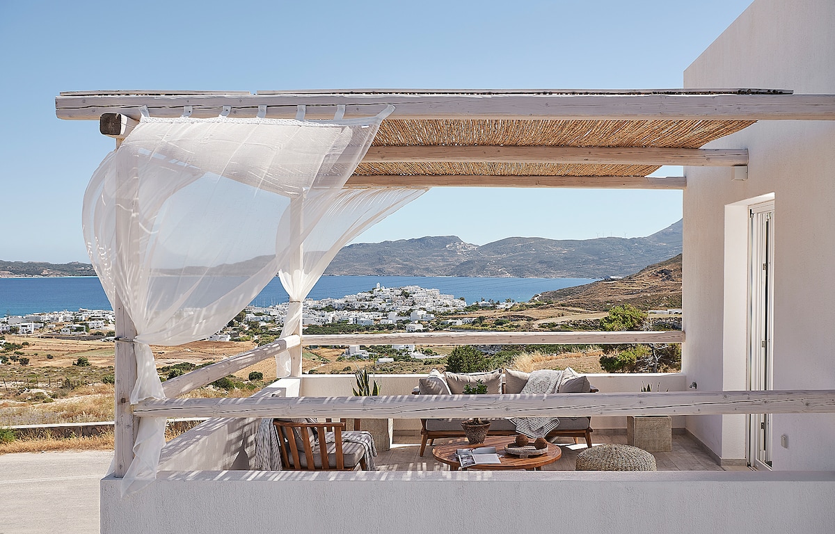 A private balcony is featured, complete with light fabric draping and wooden furniture. A coffee table is centrally placed, surrounded by seating that offers scenic views of the landscape and sea. The serene coastal surroundings add to the inviting atmosphere.