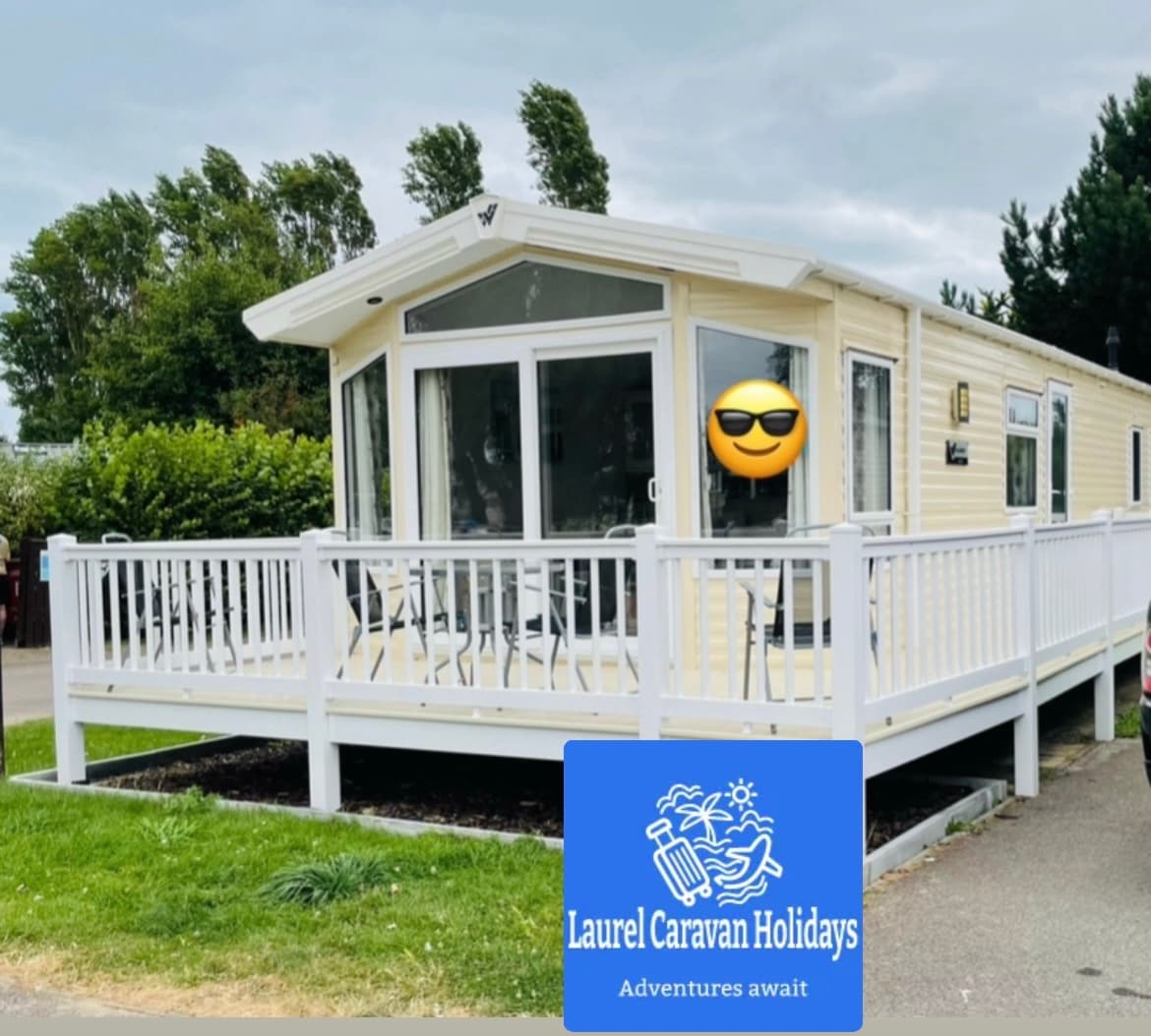 A light yellow caravan features large windows and a wrap-around deck, providing ample outdoor seating. The deck is enclosed with a white railing, and the surrounding greenery creates a peaceful ambiance. A logo for Laurel Caravan Holidays is visible at the bottom of the image.