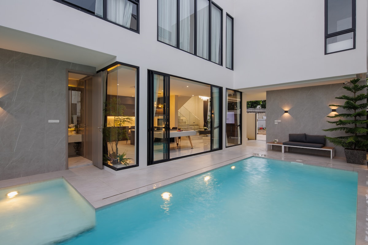 A contemporary villa features a spacious indoor and outdoor layout, with large glass doors providing easy access to a serene swimming pool. Soft lighting illuminates the water, while a comfortable seating area is positioned nearby, surrounded by greenery.