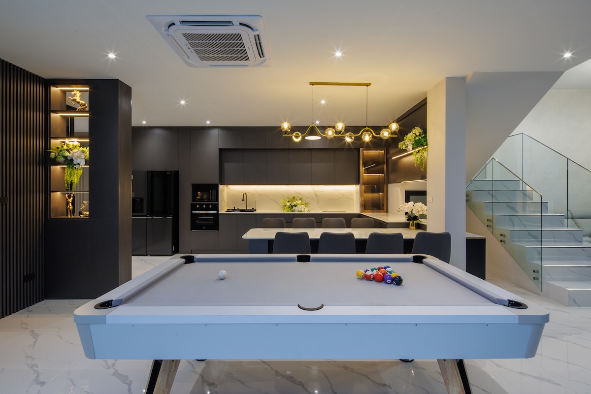An elegant game room features a white pool table with colorful balls arranged at one end. Modern cabinetry in dark tones contrasts with the light marble flooring. Ambient lighting highlights the kitchen area in the background, showcasing sleek appliances and contemporary decor.
