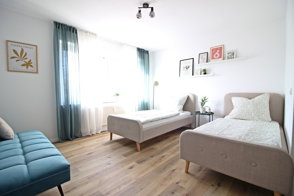The room features two single beds positioned with ample space between them. A light-toned rug complements the wood floor. Natural light floods in through a large window, adorned with sheer curtains. A cozy couch and minimalist shelves with framed artwork add a modern touch.