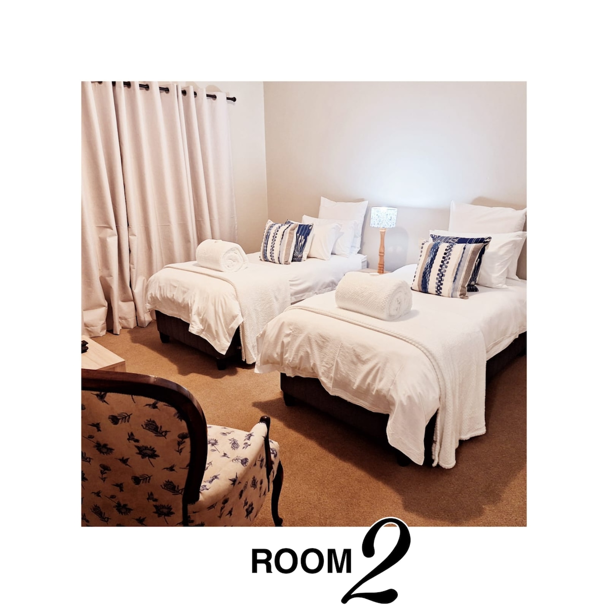 A serene bedroom is featured with two spacious beds, dressed in crisp white linens. Decorative pillows add a touch of color. A small bedside lamp provides soft lighting, while light curtains frame a window, allowing subtle light to enter the room.