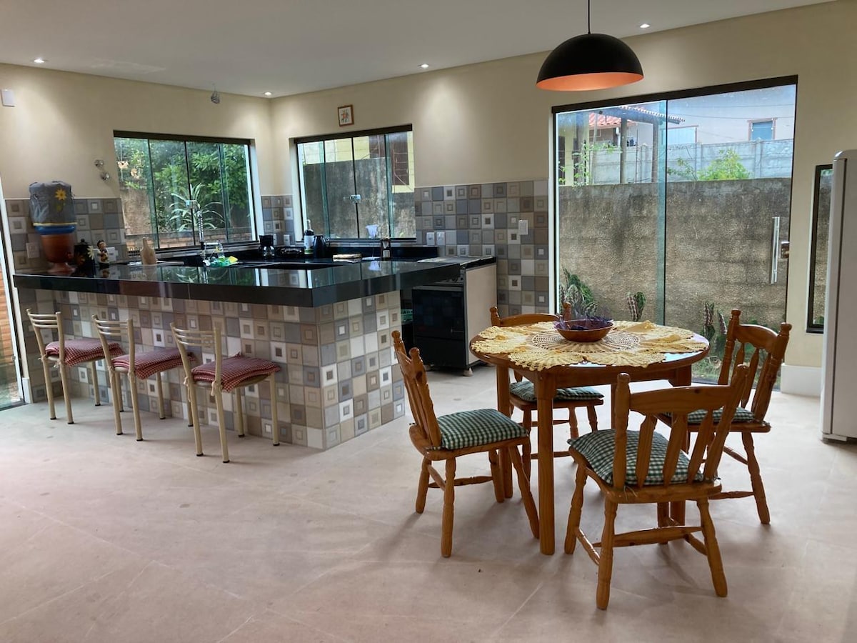 A spacious kitchen area features a central island with a dark countertop, complemented by patterned wall tiles. A round wooden table with four chairs is positioned near large windows, allowing natural light to illuminate the space. Appliances include a refrigerator and an oven.