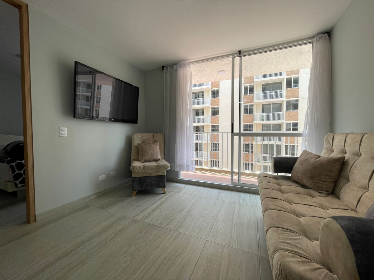 A minimalist living area is presented with a neutral color palette, featuring a modern couch and an armchair. Large floor-to-ceiling windows allow natural light to fill the space, with a view of the building's exterior visible through the glass.