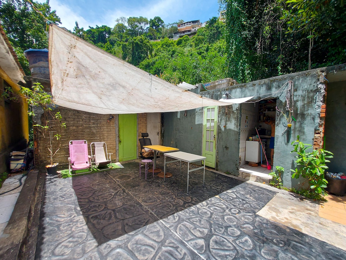 An outdoor space is presented, featuring a textured floor and a shaded area created by a large tarp. Two chairs and a small table are arranged for seating, with green walls and plants visible in the background. Natural greenery frames the area, enhancing a connection to the surroundings.