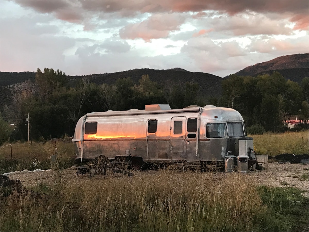 An Airstream trailer rests on a gravel bed amidst tall grasses, reflecting the warm colors of the sunset. Surrounding trees and an expansive sky filled with clouds provide a natural backdrop, while Jicarita Peak is visible in the distance.