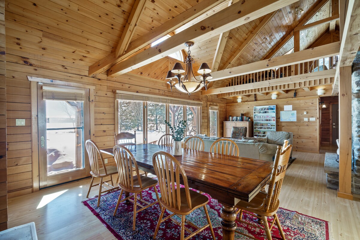 The spacious dining area features a wooden table that comfortably seats eight, illuminated by a chandelier. Rich wooden beams and walls create a rustic ambiance, while large windows provide natural light and views of the surrounding nature.