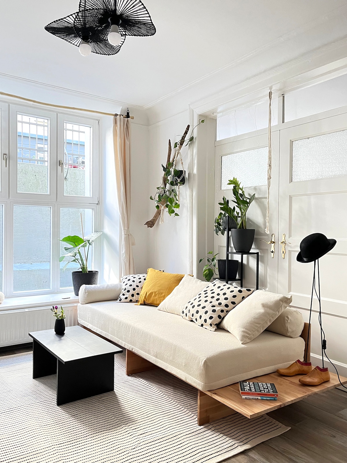 Sebule ya kisasa yenye sofa ya rangi ya mchanga na madirisha makubwa ambayo huingiza mwanga mwingi wa asili. Mapazia angavu, kona maridadi ya kijani kibichi, meza ya kahawa ya kifahari na taa ya kisanii huunda mazingira mazuri ya kisasa.