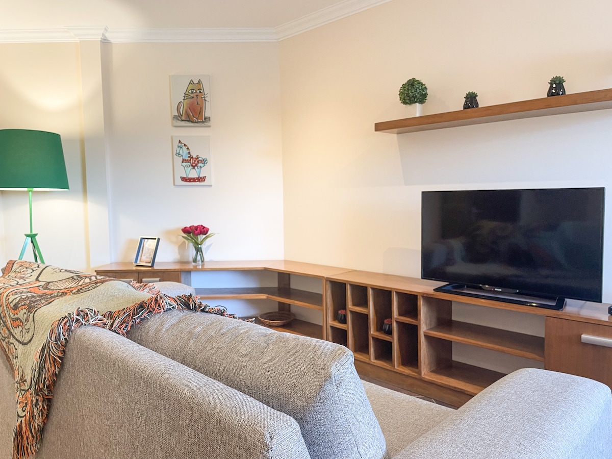 A living area features a light-colored sofa with a decorative blanket draped over it. A wooden entertainment unit holds a television and a few decorative items. Wall art with nature themes is displayed above the unit, complemented by a potted plant on the shelf.