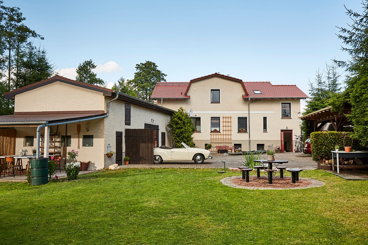 A spacious outdoor area is featured, showcasing two buildings with a mix of rustic and modern architecture. A classic car is parked in front of the main structure. Around a small fire pit, several chairs are arranged, surrounded by manicured green grass.