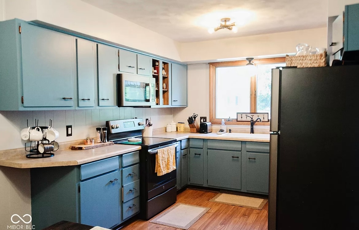 A well-equipped kitchen features blue cabinetry and light-colored countertops. Appliances include a refrigerator, stove, and microwave. Natural light enters through a large window above the sink, and a coffee maker is positioned near the counter for convenience.