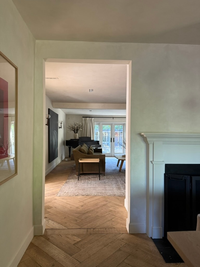 A view through an open doorway highlights a spacious living area with a piano and comfortable seating. Natural light filters through large glass doors, leading to the exterior. The hardwood floor features a herringbone pattern, contributing to a warm atmosphere.