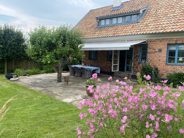 An outdoor patio area is surrounded by a well-maintained garden featuring colorful flowers and lush grass. A large shaded table with wicker chairs is positioned next to a tree, providing a relaxing spot to enjoy the scenery. The brick house has blue-framed windows.