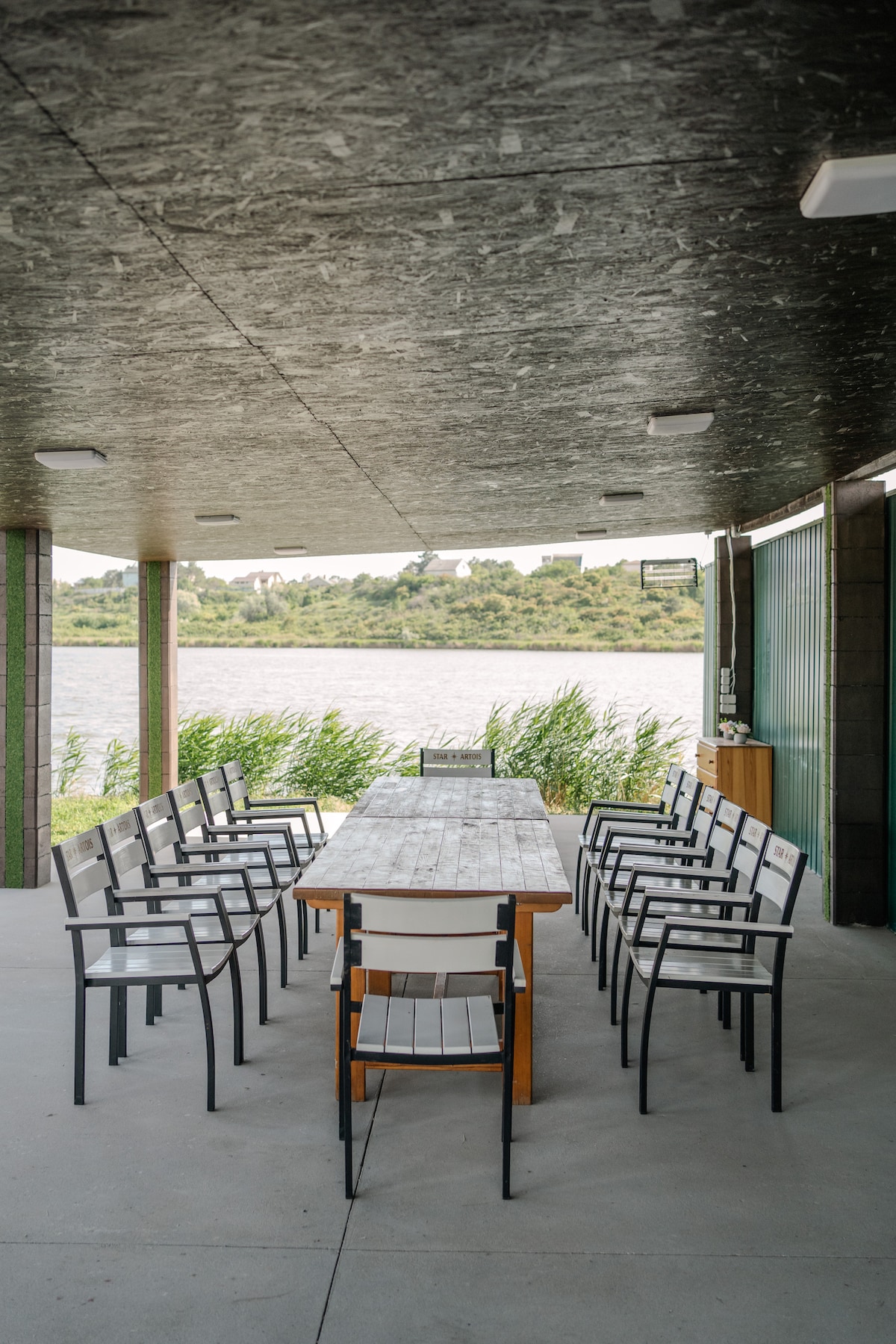 A spacious outdoor dining area features a long wooden table surrounded by twelve chairs under a covered terrace. The serene view of the water is complemented by lush greenery in the background, creating an inviting space for gatherings.