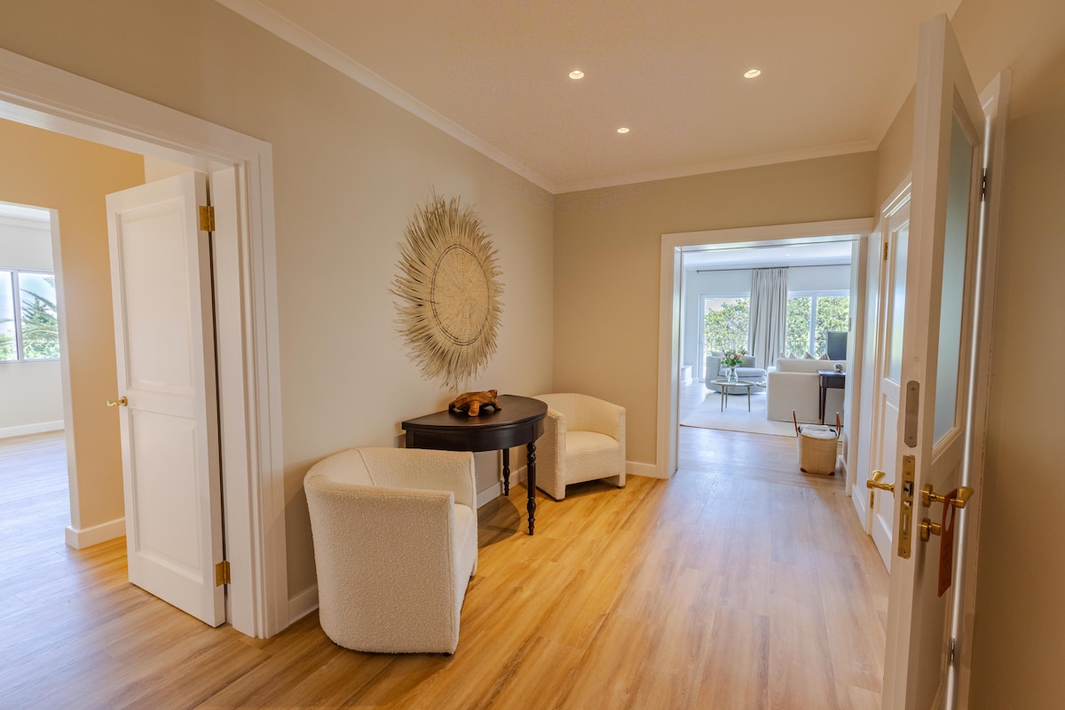 A well-lit hallway features light-colored walls and hardwood flooring. A round mirror adorns the wall, adding depth, while a small table is set with decorative items. White armchairs provide seating, creating a welcoming transition to the adjoining living area.