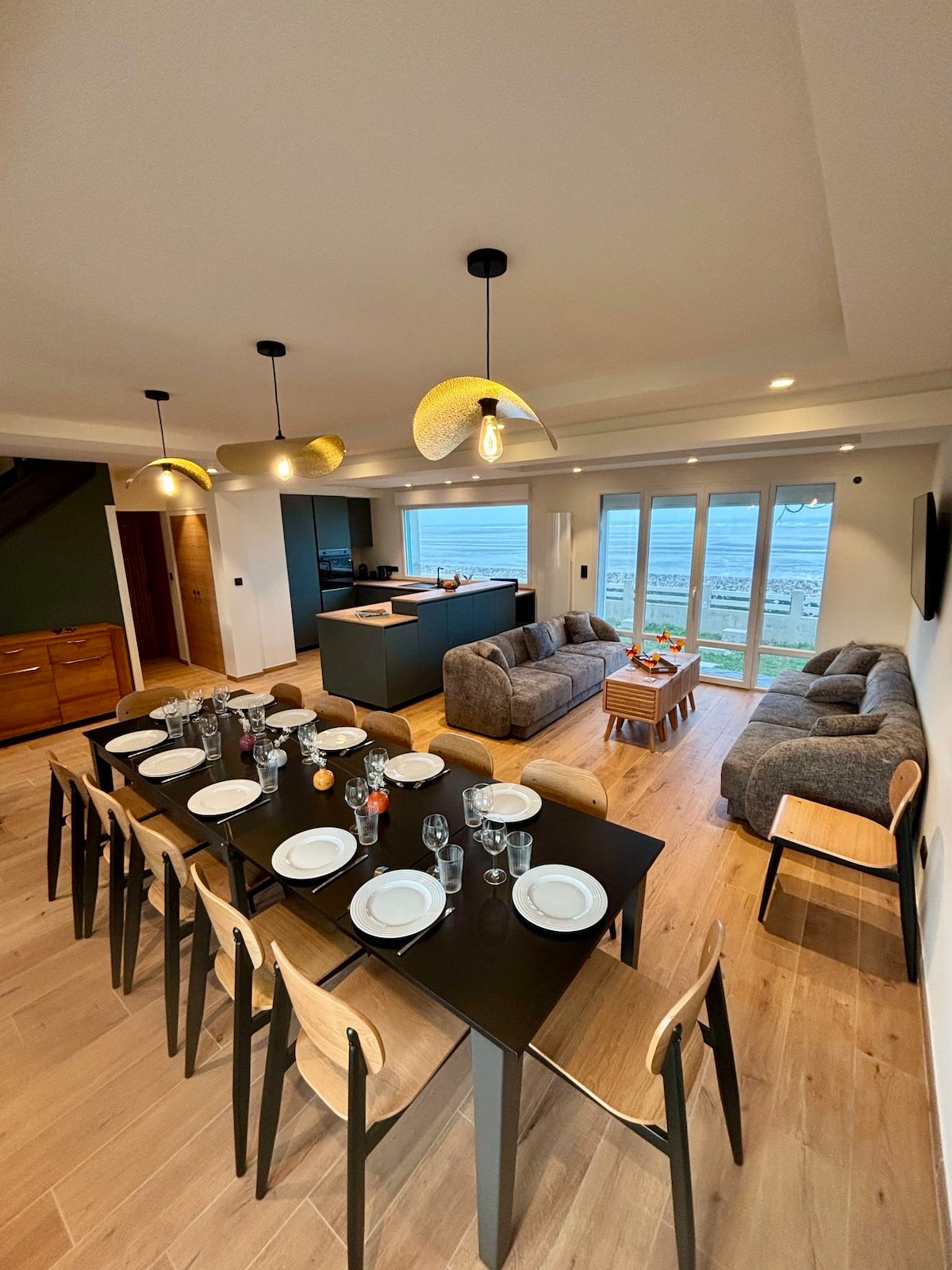An open-concept living area features a dining table set for eight, complemented by modern chairs. A cozy seating arrangement is visible, with a light grey sofa facing expansive glass doors that provide views of the beach. Soft lighting from stylish fixtures adds warmth to the space.