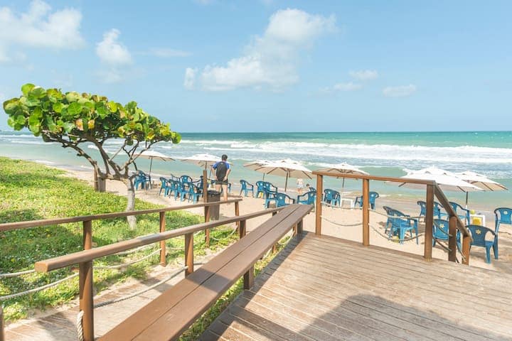 A wooden deck leads to a sandy beach, with umbrellas and blue chairs arranged for guests. A tree with broad leaves provides shade nearby, while gentle waves roll onto the shore under a clear sky.