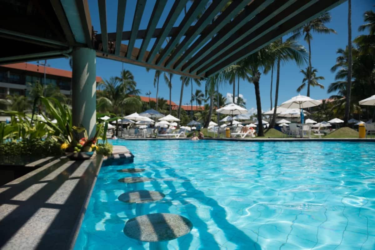 A clear swimming pool is framed by wooden slats above, providing shade. Surrounding lounge chairs and umbrellas are visible, with palm trees in the background. The vibrant blue water reflects sunlight, creating a relaxing ambiance within the resort's outdoor recreational area.