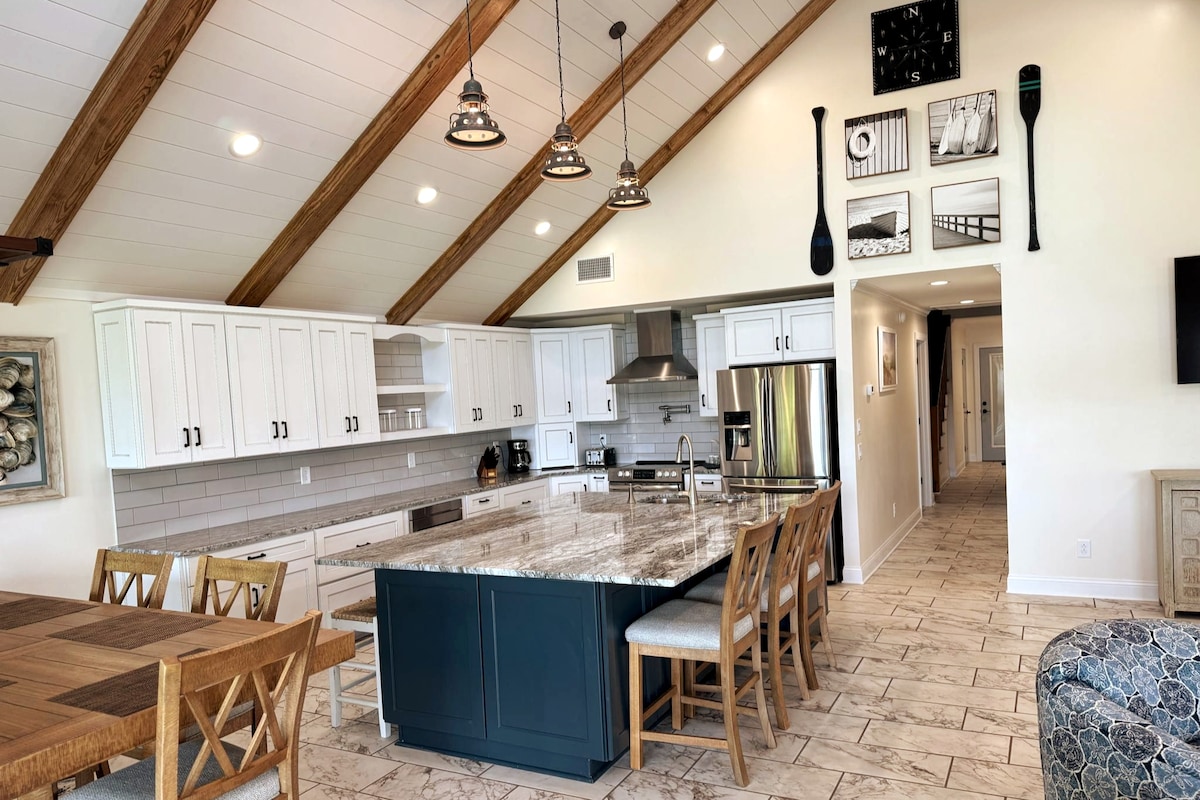 An open-concept kitchen features white cabinetry and a large island with marble countertops. High wooden beams accentuate the ceiling, while modern pendant lights hang above the island. A dining area is visible, and nautical decorations adorn the walls, enhancing the coastal theme.