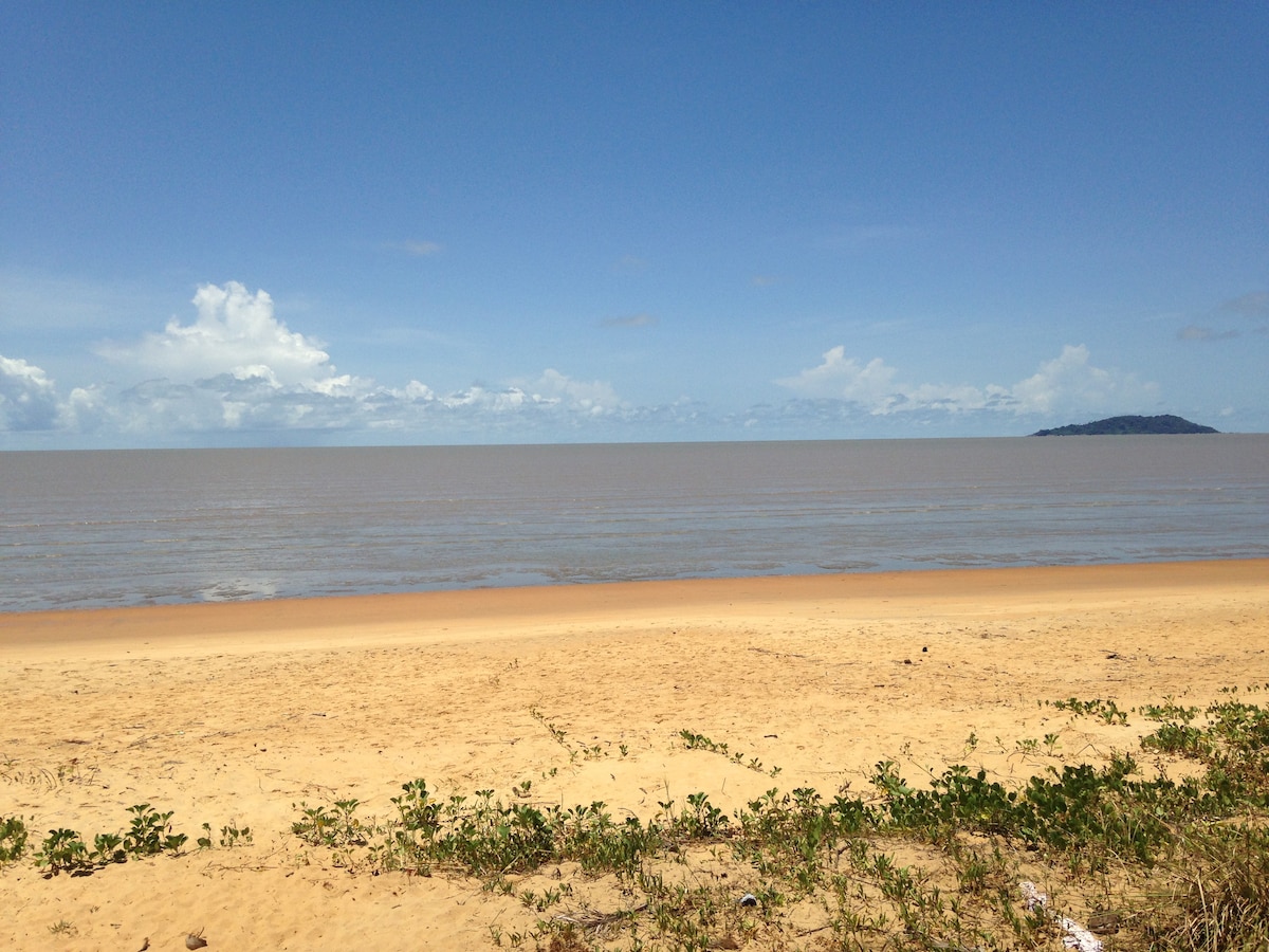 A serene beach landscape features soft golden sand stretching toward the calm water. Gentle waves lap at the shoreline, and a distant island is visible on the horizon. The sky above is clear with a few fluffy clouds, enhancing the tranquil atmosphere.