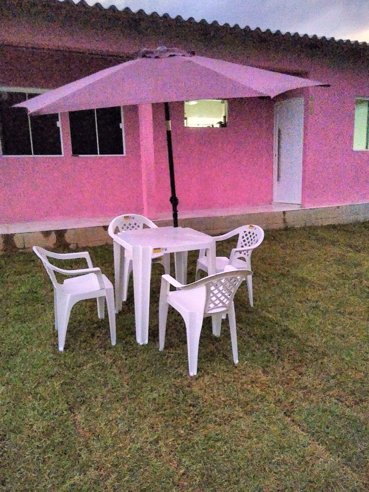 An outdoor seating area is presented, featuring a white table accompanied by four matching chairs. A large pink umbrella provides shade above the table, situated on a grassy lawn. A pink building with multiple windows serves as the backdrop.