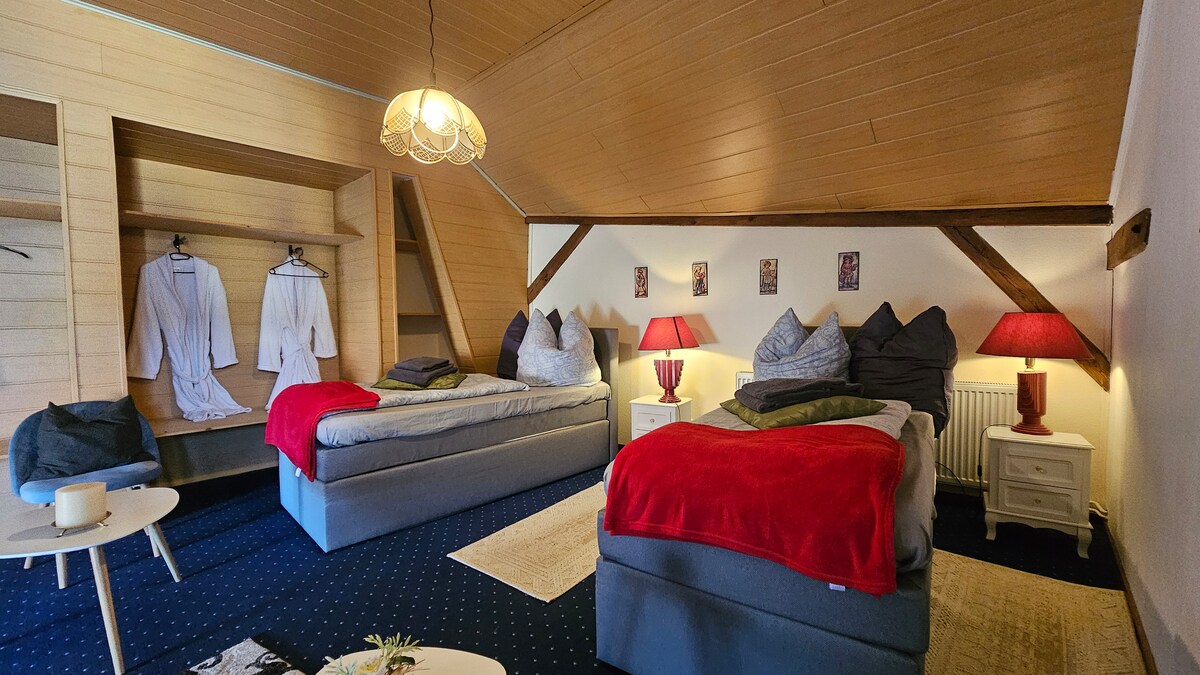 The room features two spacious beds, each adorned with a set of pillows and warm blankets. A cozy seating area is present with a small side table. Soft lighting highlights the wooden beams and walls, complemented by bathrobes hung nearby.