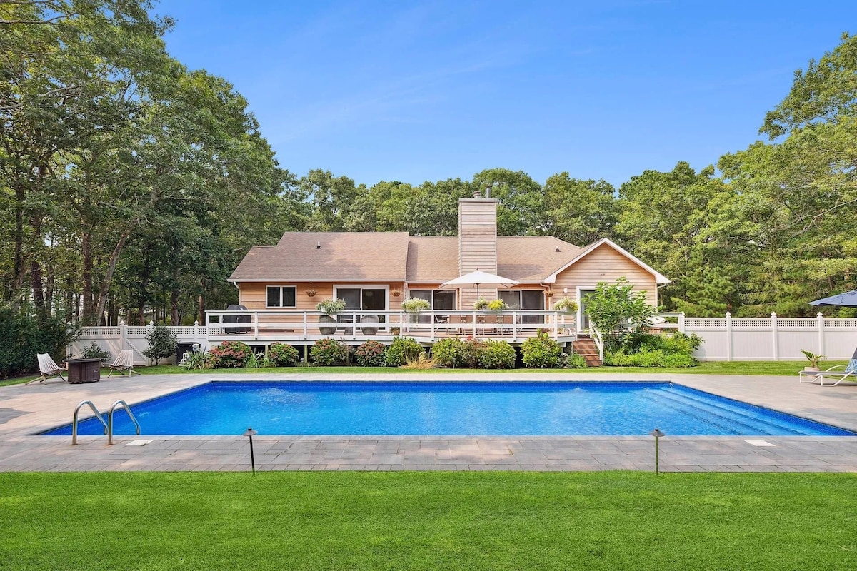A spacious outdoor area features a large saltwater pool surrounded by well-maintained grass. The two-story house is seen in the background, with a deck that includes plants and provides a tranquil setting. Trees border the property, offering a sense of privacy.