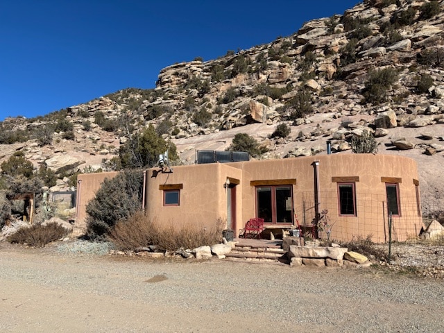 A single-story casita is nestled against a rocky hillside, featuring adobe-style architecture. Large windows and a welcoming entrance invite natural light, while a small patio area with seating is visible in front. Surrounding vegetation includes native shrubs and rocky terrain, enhancing the natural setting.