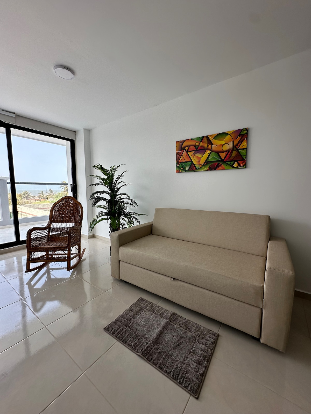 A light-filled seating area features a beige sofa and a woven chair beside a potted plant. A piece of abstract art adds color to the wall, while large glass doors provide views of the outdoors. A small mat is placed on the glossy tiled floor.